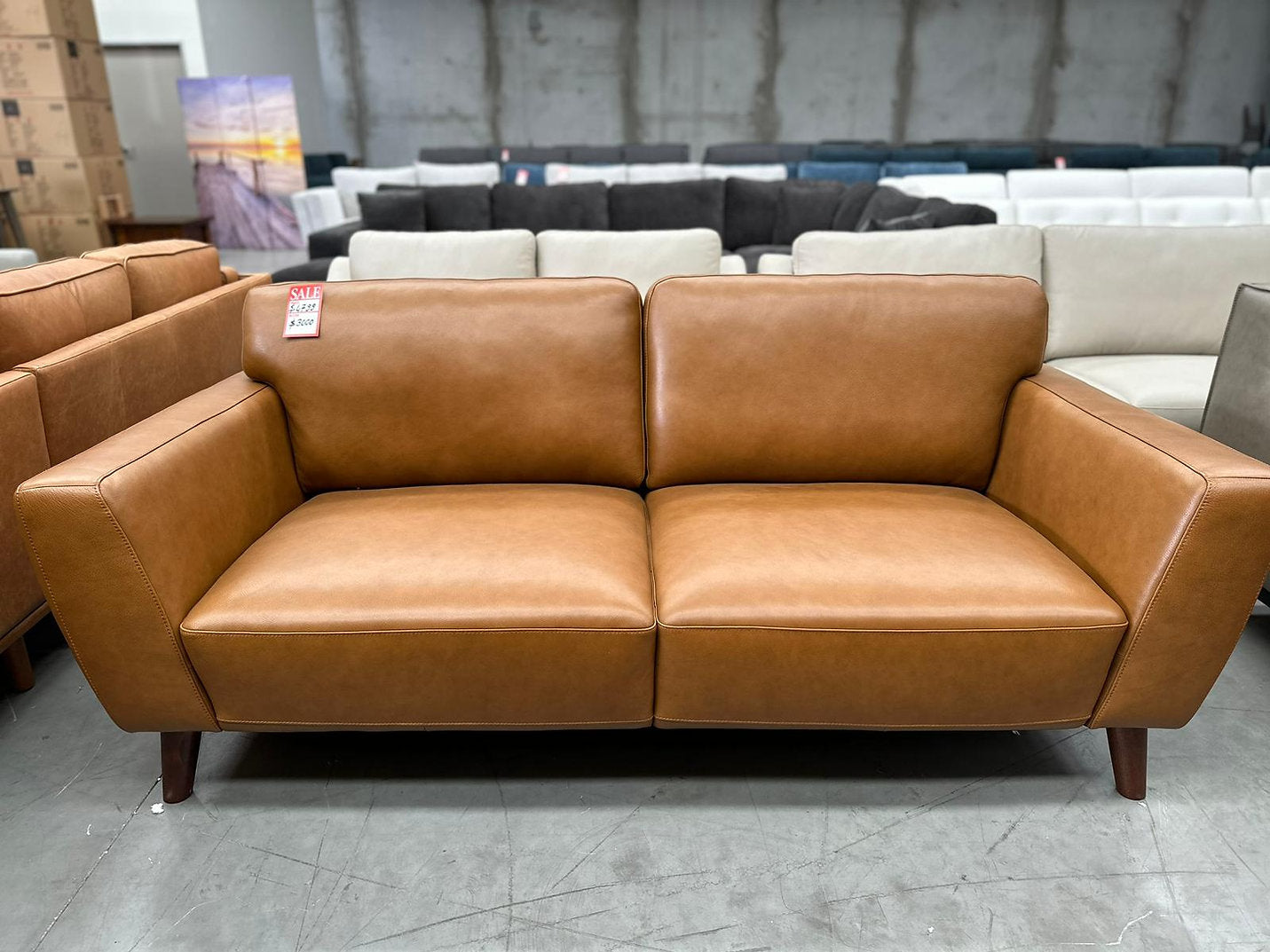 Brown leather sofa in a showroom setting with other furniture in the background.